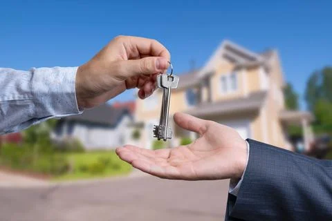 A hand giving a key to another hand. House in the background. Stock Photos