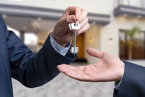 A hand giving a key to another hand. House in the background. Stock Photos