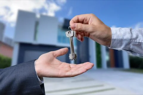 A hand giving a key to another hand. House in the background. Stock Photos