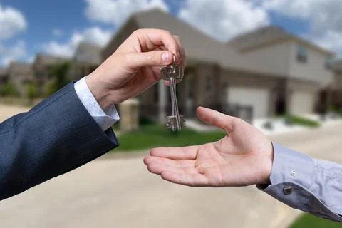 A hand giving a key to another hand. House in the background. Stock Photos