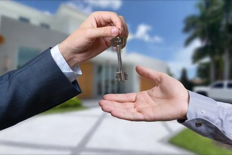 A hand giving a key to another hand. House in the background. Stock Photos