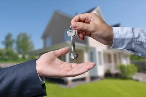 A hand giving a key to another hand. House in the background. Stock Photos