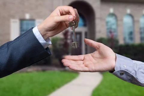A hand giving a key to another hand. House in the background. Stock Photos