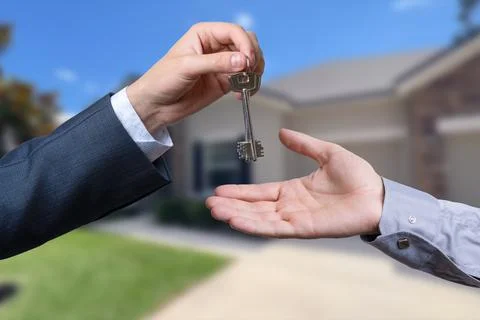A hand giving a key to another hand. House in the background. Stock Photos