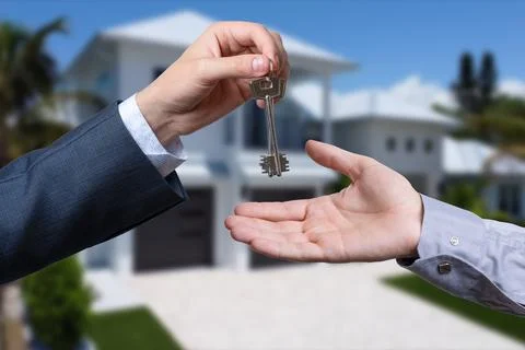 A hand giving a key to another hand. House in the background. Stock Photos