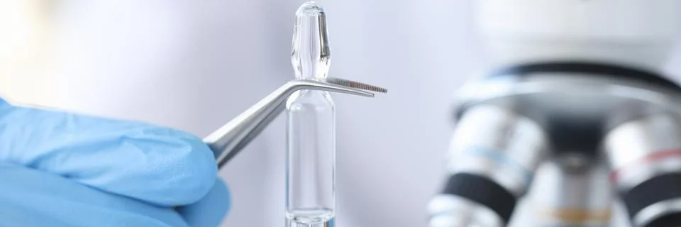 Hand in glove holds vial with vaccine against background of microscope Stock Photos