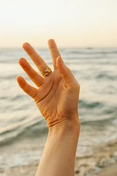 A hand with a gold ring points towards the ocean. The sun is setting in the b Stock-Fotos