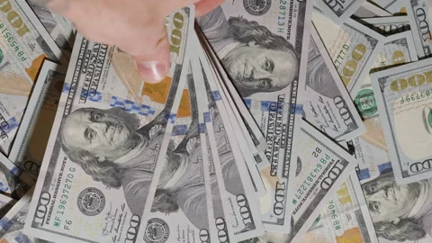 A hand grabs a stack of hundred-dollar bills from a scattered pile, lifting them Stock Footage 296174491
