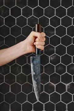 A hand gripping a sharp chef's knife against a textured hexagonal tile back.. Stock Photos