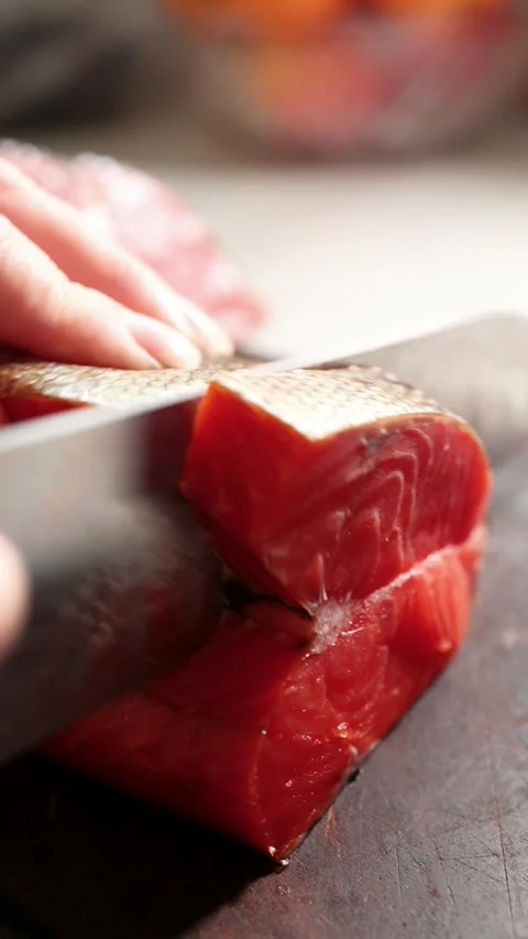 Hand guided cleaver slicing thick raw salmon loin on dark cutting board in Stock Footage 339914982