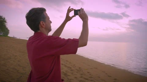 Hand held shot of a middle aged man taking photos of a sunrise on the beach Stock Footage 54624343