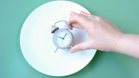 Hand holding alarm clock against empty plate, intermittent fasting concept. Stock Footage 294492802