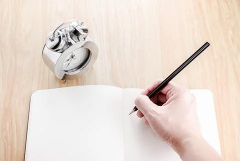 Hand holding black pencil and writing on blank open notebook with alarm clock Stock Photos
