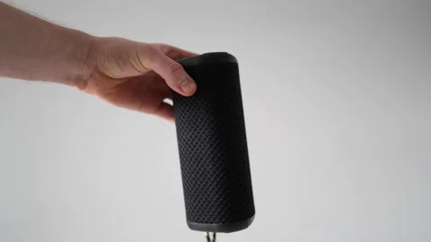 A hand holding a black portable Bluetooth speaker with a textured surface and a  Stock Footage 303908257