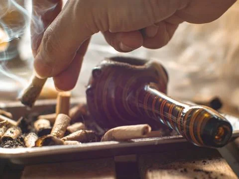 Hand holding a burning joint roach with rising smoke above an ashtray full .. Stock Photos
