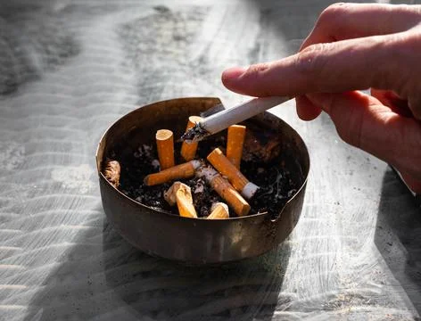 Hand holding a cigarette whose ashes are tapped into an ashtray Stock Photos