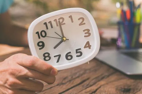 The hand holding the clock is at the work desk. Stock Photos