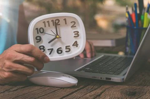 The hand holding the clock is at the work desk. Stock Photos