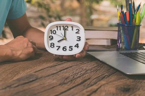 The hand holding the clock is at the work desk. Stock Photos