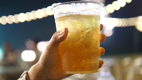 Hand holding a cold beer in a plastic cup at a night market with bokeh lights Stock Footage 309978832