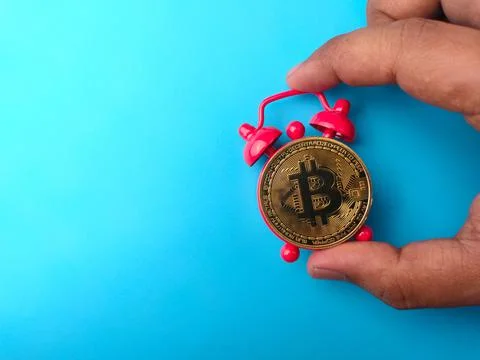 Hand holding colored alarm clock with golden bitcoin on a blue background wit Stock Photos