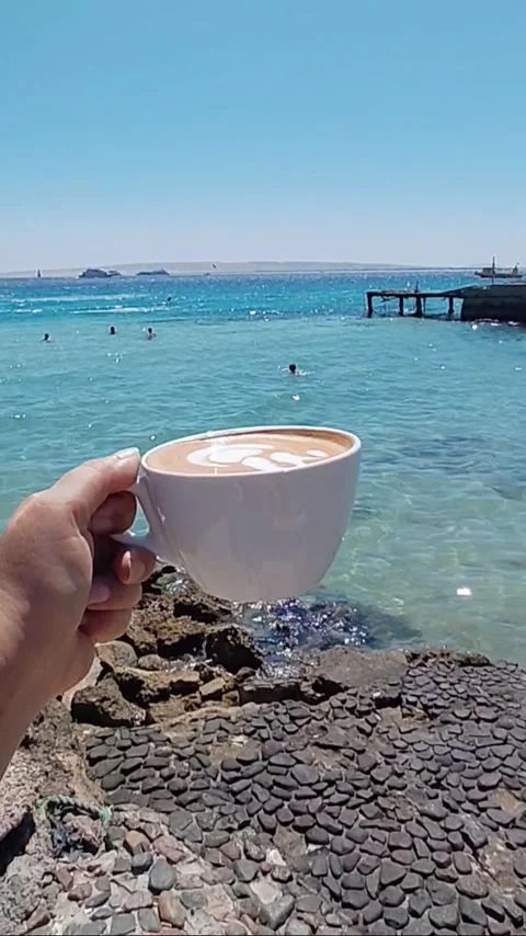 Hand holding cup of latte coffee against turquoise sea and beach resort Stock Footage 340014421