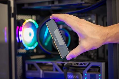 Hand holding DDR5 RAM module in front of gaming computer with RGB lighting,.. Stock Photos