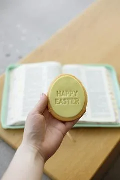 Hand holding Easter cookie with greeting over open Bible at home, copy space. Stock Photos