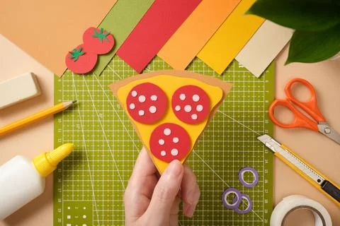 Hand is holding a finished paper craft: a slice of pepperoni pizza. There are Stock Photos