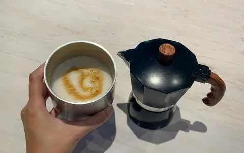 Hand Holding Foamed Milk Coffee Beside Black Moka Pot Stock Photos