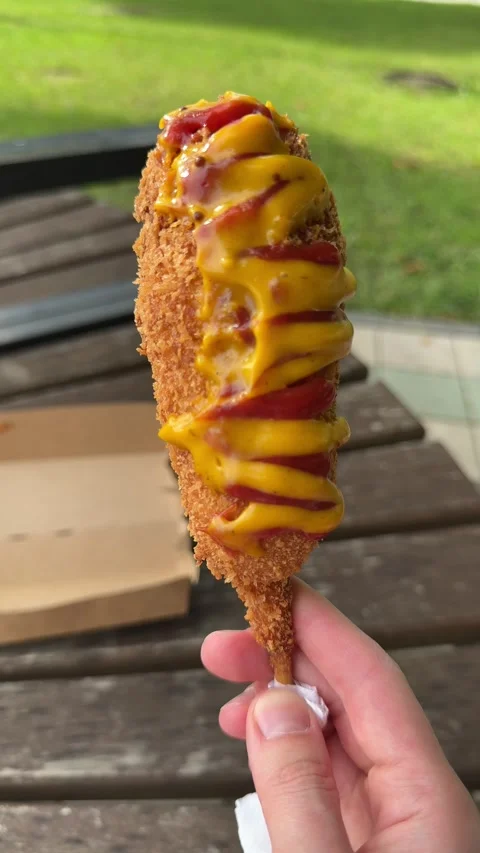 Hand holding fried corn dog with mustard and ketchup on wooden stick 스톡 동영상 338361782