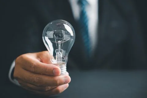 A hand holding a lightbulb with a blurred background of a man in a suit. Stock Photos