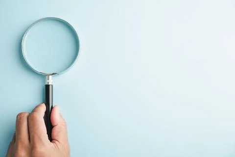 A hand holding a magnifying glass on a light blue background. Stock Photos