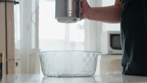 Hand holding metal sifter tilted above empty glass mixing bowl in bright kitchen Stock Footage 343675725