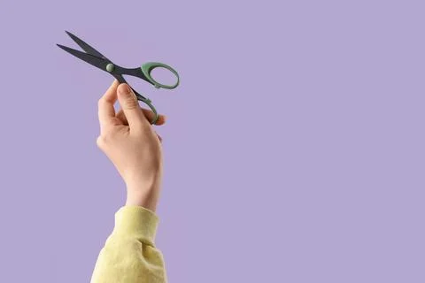 Hand holding open scissors with green handles on a solid purple background, c Stock Photos