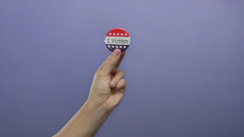 Hand holding a patriotic button reading 'i voted' against a solid purple ba.. Stock Footage 312860125