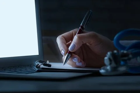 Hand holding pen writing notes next to laptop and stethoscope in dim light Stock Photos