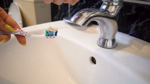 Hand holding a pink toothbrush with toothpaste on top of sink under a running Stock Footage 102352454