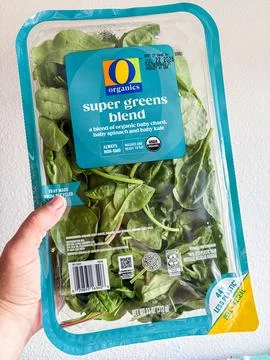 A hand holding a plastic container of O Organics Super Greens Blend salad mix Stock Photos