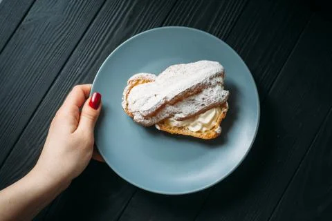 Hand holding a plate with eclair. Eclair with half-cream under powdered sugar Stock Photos