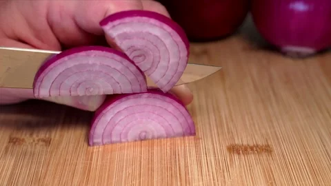 A hand is holding a red onion on a cutting board and slicing it with a knife. Stock Footage 341345786