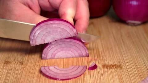 A hand is holding a red onion on a cutting board and slicing it with a knife. Stock Footage 341346020