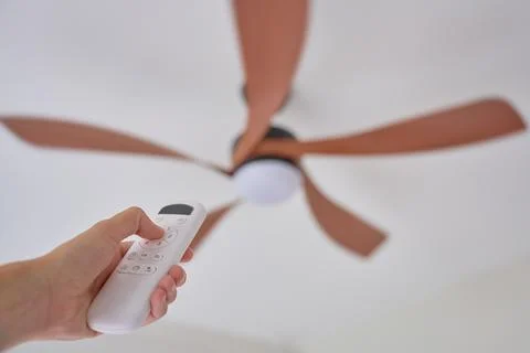 Hand holding remote control aimed at spinning ceiling fan indoors Stock Photos