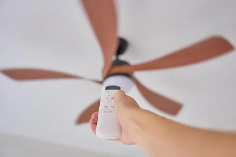 Hand holding remote control aimed at ceiling fan in home interior Stock Photos