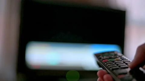 Hand Holding Remote Control Pointing At Blurred  TV Screen In Living Room Stock Footage 327957387