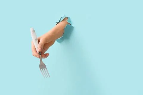 Hand holding silver fork with white marble handle through torn blue paper bac Stock Photos