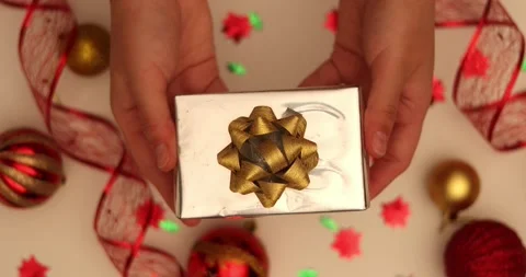 Hand holding a silver gift box with a gold bow surrounded by Christmas ornaments Stock Footage 339071449
