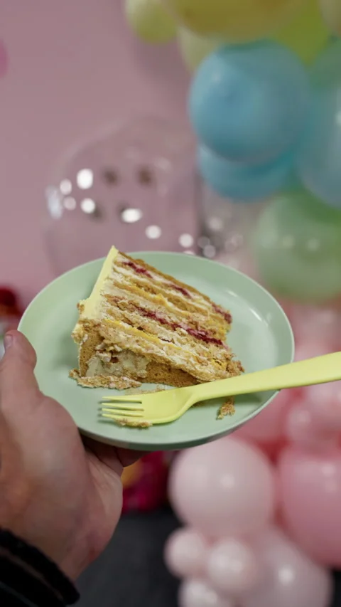Hand holding slice of layered birthday cake on plate with fork and soft balloon Stock Footage 345605762