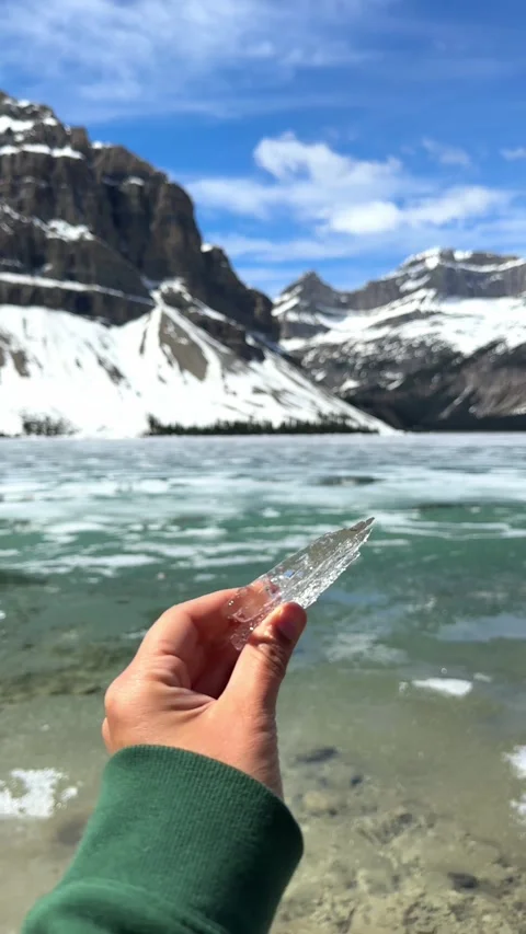 Hand holding a small piece of clear ice with a frozen mountain lake and snowy Vidéo 333869036