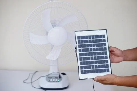 Hand holding small solar panel in front of electric fan at home. Renewable energ Stock Photos